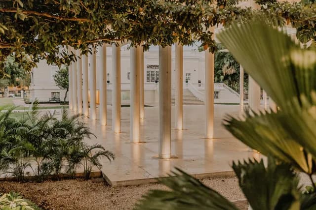Columnata del pabellón iluminada al atardecer entre palmeras, con la escalinata de la casona al fondo