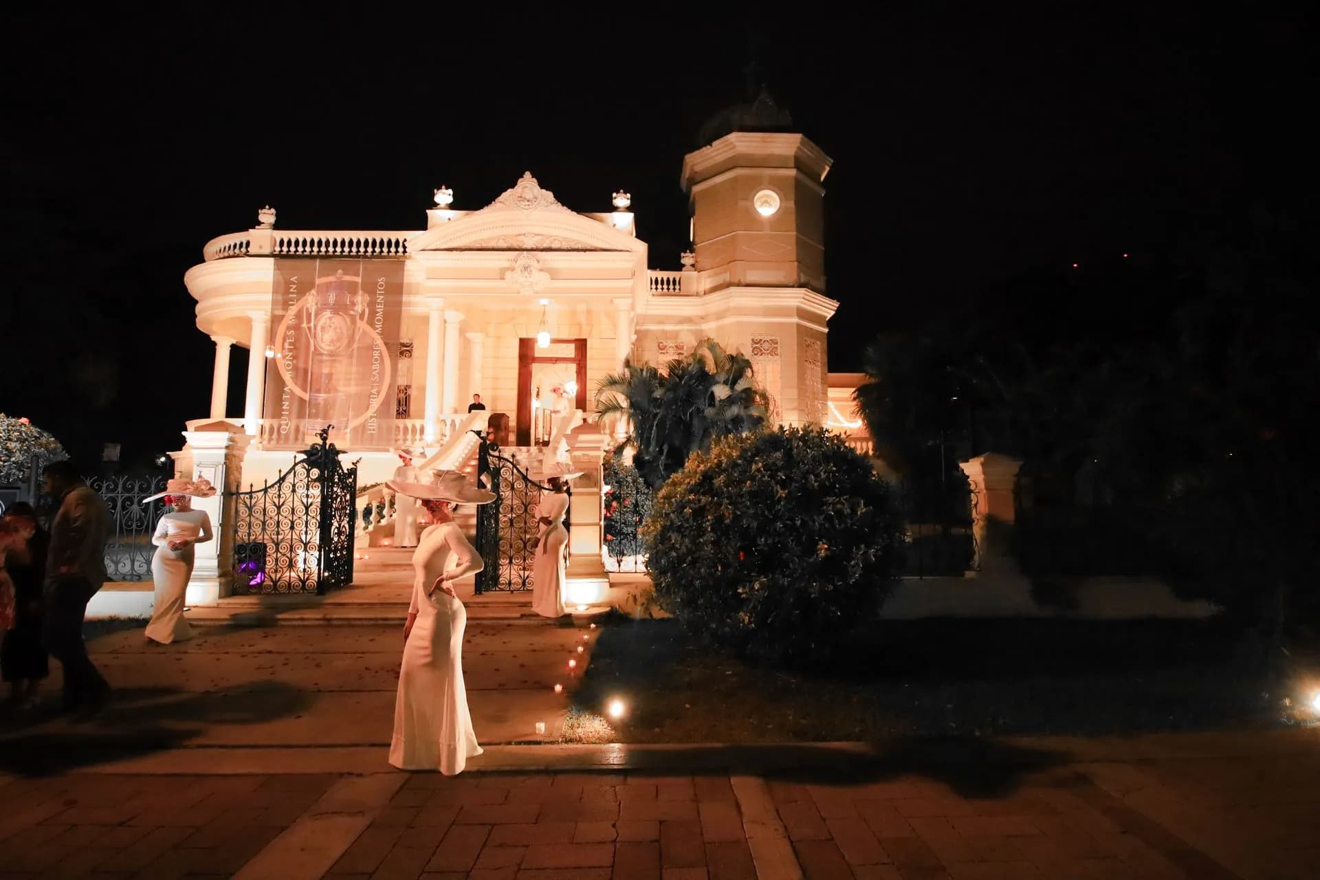 La casona iluminada de noche con actrices en vestidos de época blancos y pamelas entre velas encendidas