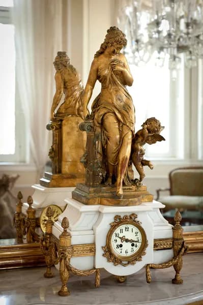 Reloj de mármol blanco con figura femenina clásica y querubín en bronce dorado, reflejado bajo un candil de cristal